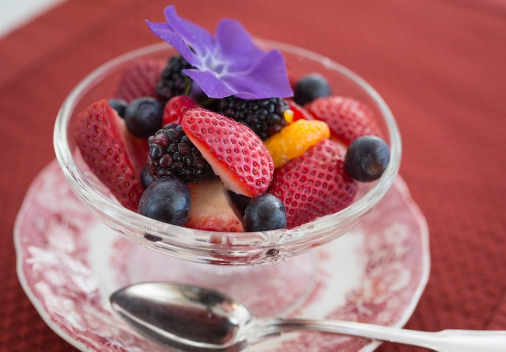 bowl of fruit with purple flower on top and spoon on red placement