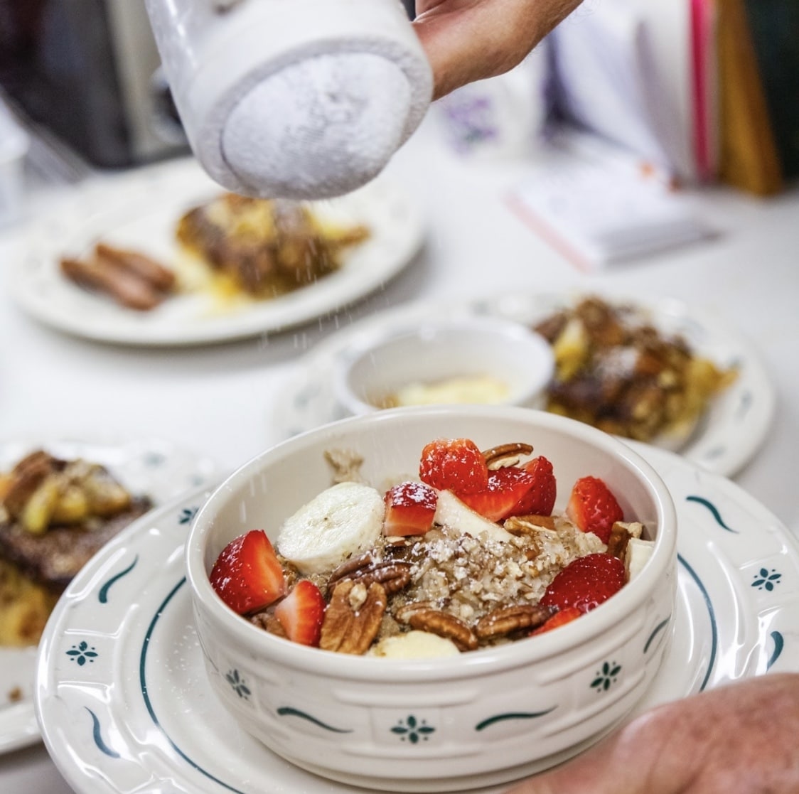 A close-up photo of a person sprinkling powdered sugar over a bowl of oatmeal. The bowl also contains sliced strawberries, bananas, and pecans.