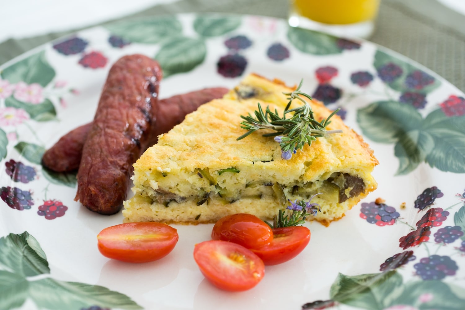 A plate with a slice of savory quiche, garnished with rosemary sprigs. Accompanying the quiche are grilled sausages and cherry tomatoes. The plate has a colorful floral pattern.