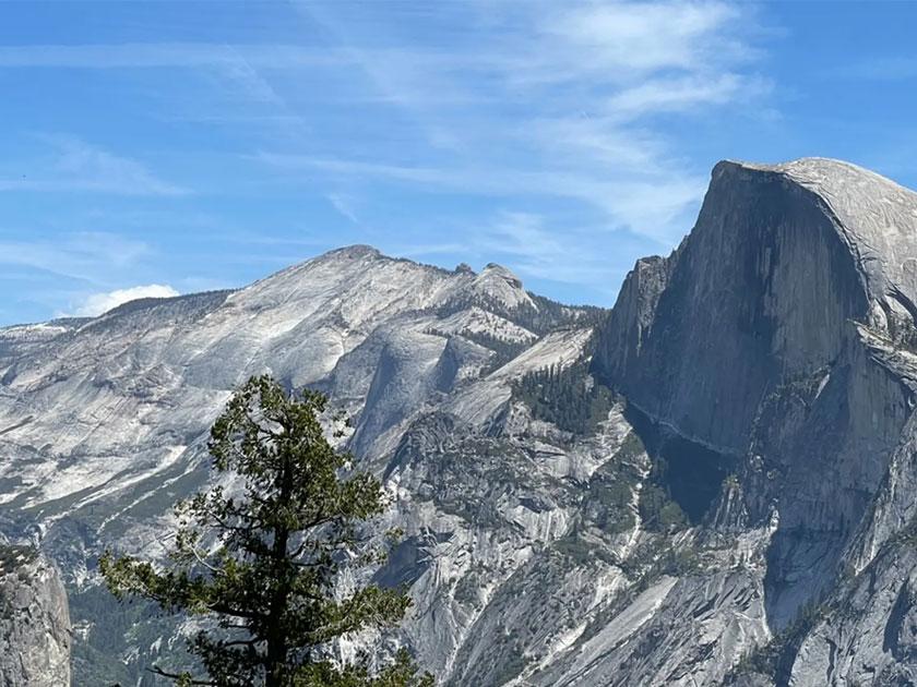 The image shows a majestic view of Half Dome, a famous granite rock formation in Yosemite National Park, with clear blue skies above and rugged mountain terrain in the background.