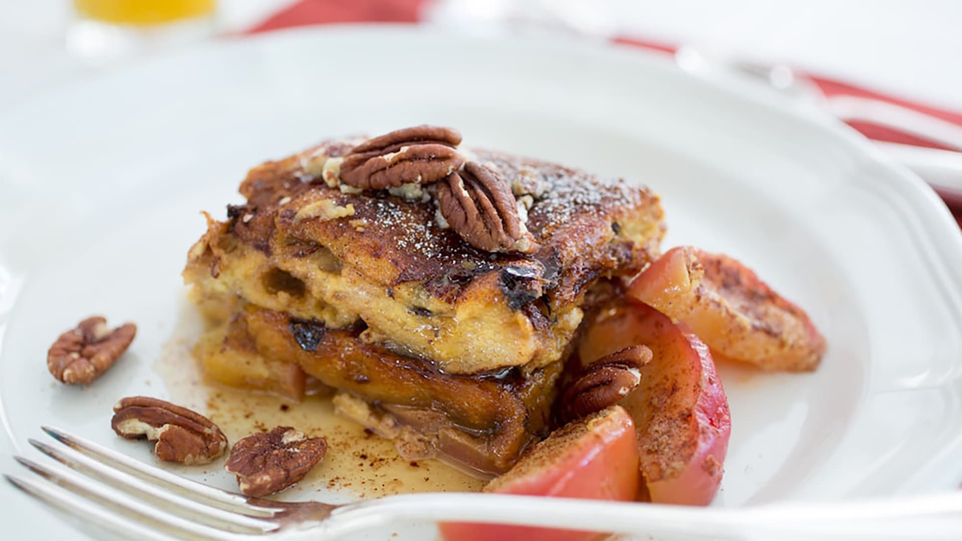 A plate of French toast topped with cinnamon and pecans. Sautéed apples are served alongside the French toast. A glass of orange juice and a fork are visible in the background.