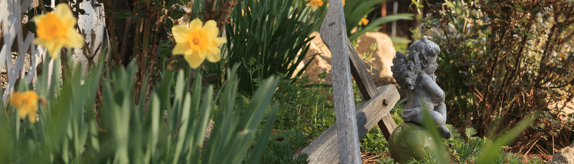 A serene garden scene with daffodils blooming in the foreground. A small, weathered statue of a cherub sits on a wooden cross, surrounded by lush greenery.