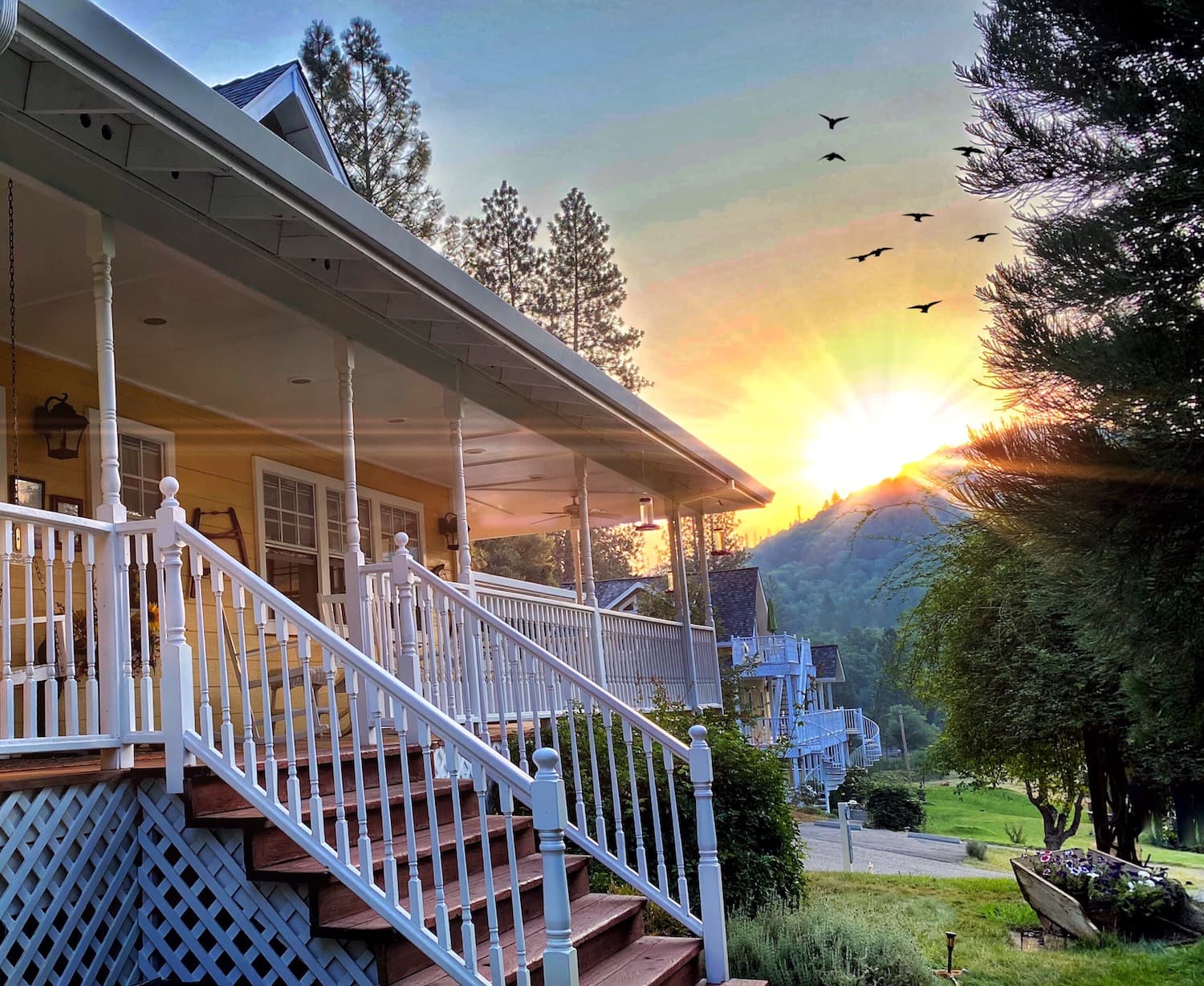 A yellow house with a white porch and stairs. The sun sets behind the house, casting a warm glow on the scene. Birds fly across the sky, and there are mountains in the distance.