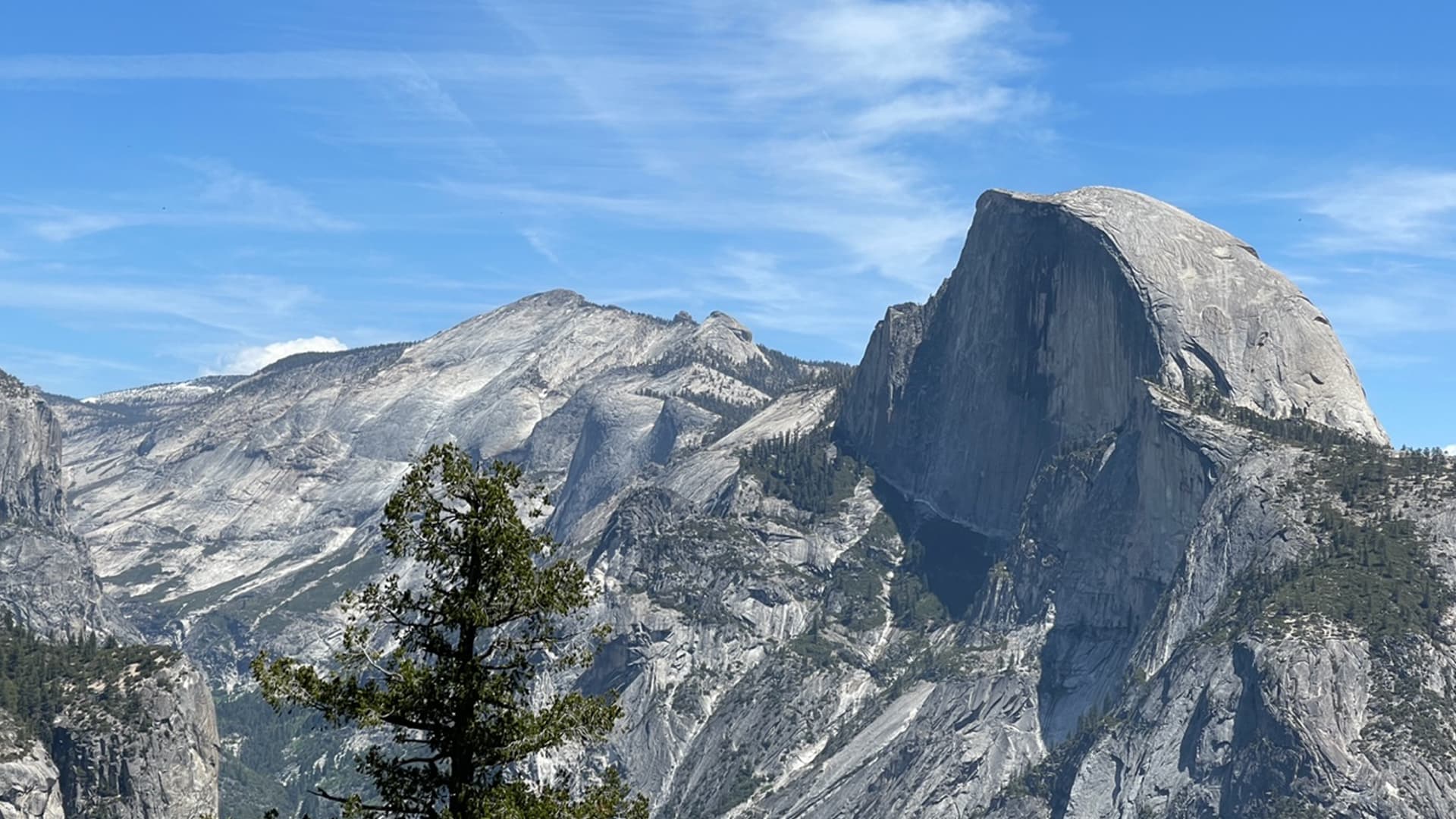 The image shows a majestic view of Half Dome, a famous granite rock formation in Yosemite National Park, with clear blue skies above and rugged mountain terrain in the background.