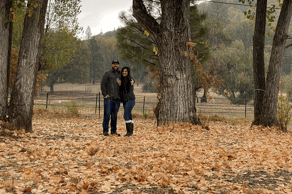 happy couple standing in fall leaves under large trees dressed for warmth