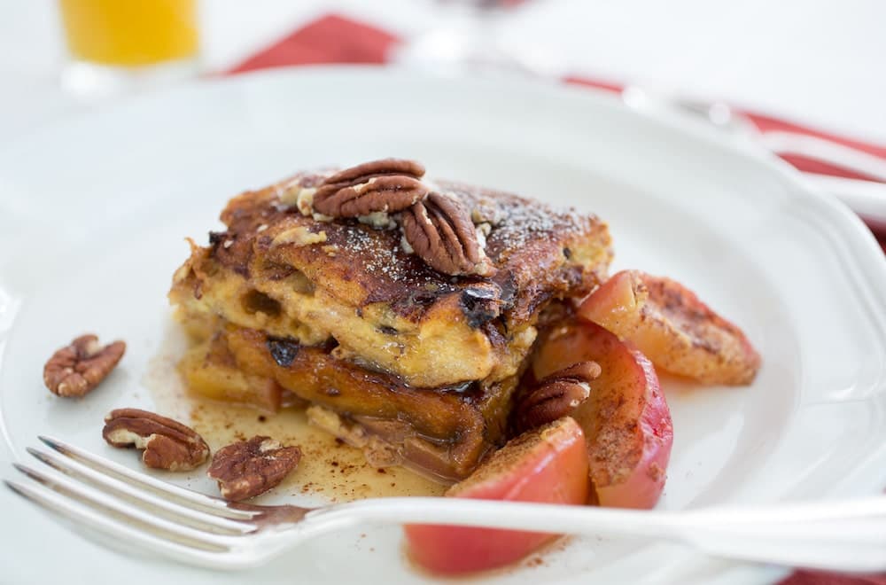 A plate of French toast topped with cinnamon and pecans. Sautéed apples are served alongside the French toast. A glass of orange juice and a fork are visible in the background.