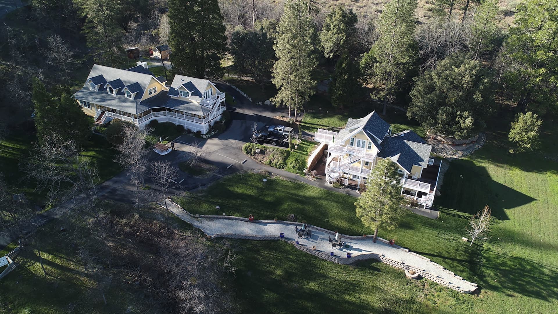 An aerial view of a secluded inn nestled in a wooded area. Multiple buildings with white balconies are visible, along with a winding pathway and a patio area with outdoor seating.