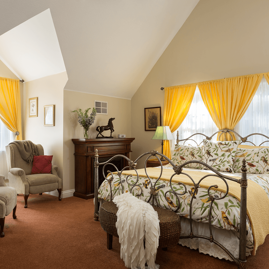 A cozy bedroom with a king-sized bed featuring a floral bedspread and yellow blanket. Two armchairs sit by a fireplace, and a painting hangs above the mantel. The room has a vaulted ceiling and teal curtains.