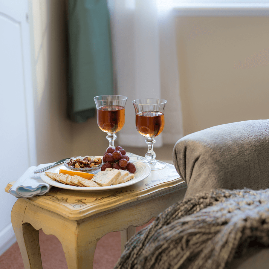 A cozy living room setting with a comfortable armchair and a small table holding two glasses of wine, a plate of cheese, crackers, and grapes. A knitted blanket is draped over the arm of the chair.