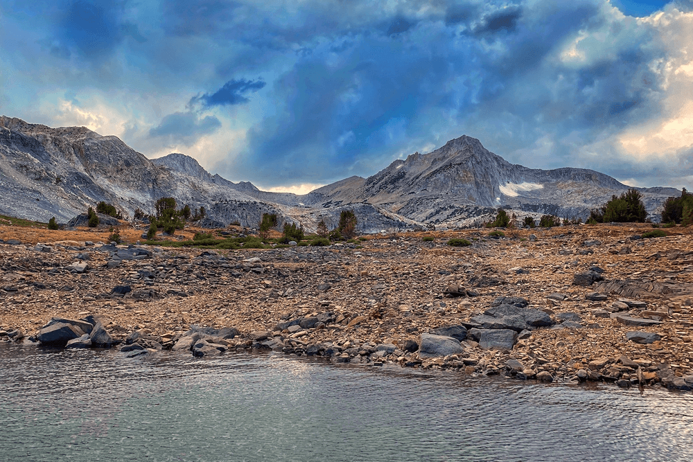 A scenic mountain landscape with a lake in the foreground. The lake level is low, exposing rocks and pebbles. Dramatic clouds fill the sky above the mountains.