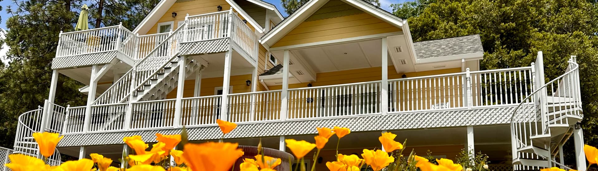 The image shows a charming yellow house with a large white-railed porch and staircase, surrounded by vibrant orange flowers in the foreground, with trees in the background under a bright sky.