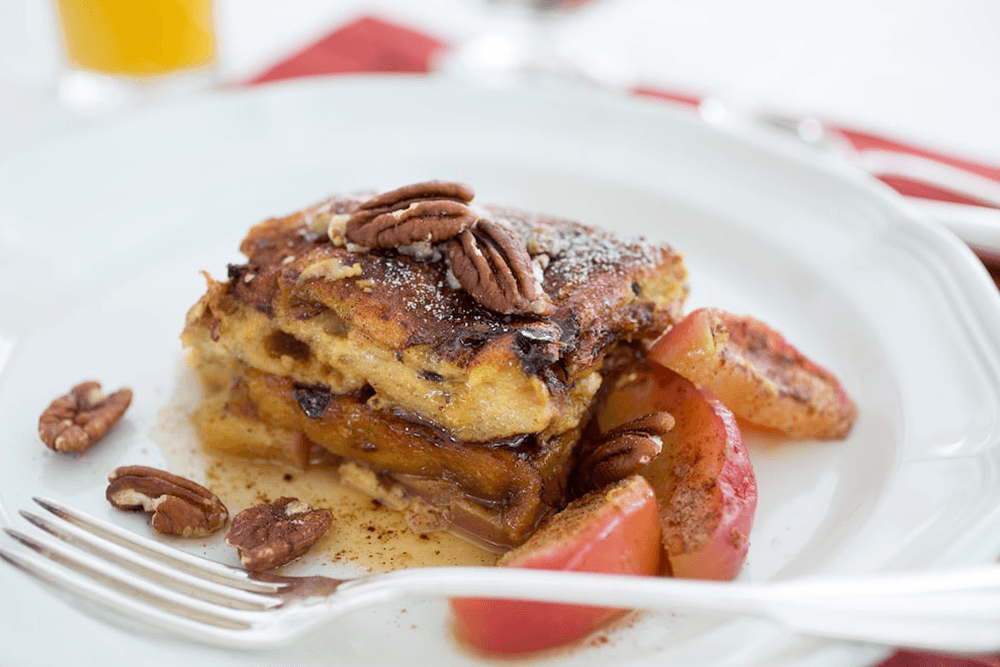 A plate of French toast topped with cinnamon and pecans. Sautéed apples are served alongside the French toast. A glass of orange juice and a fork are visible in the background.