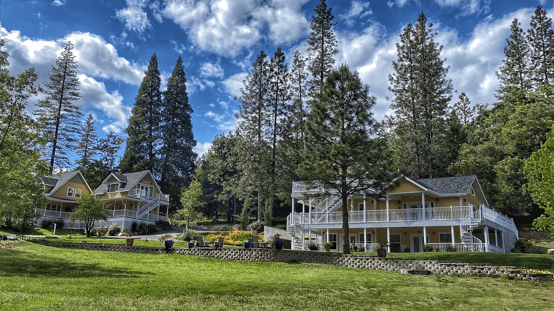 A scenic view of a yellow inn surrounded by tall pine trees. The inn has multiple buildings with white balconies and a large lawn in front. The sky is blue with fluffy white clouds.