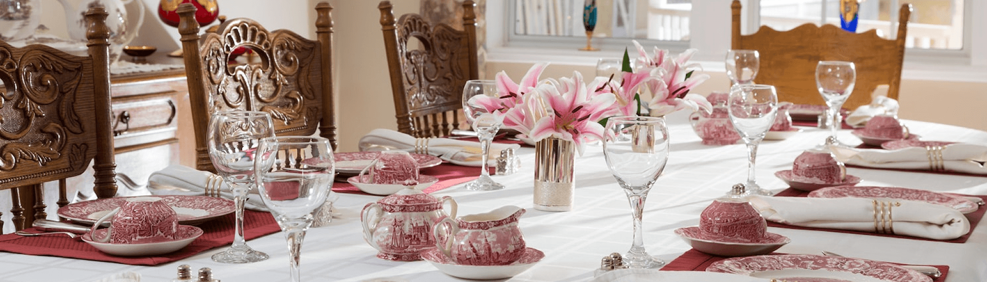 A beautifully set dining table with a white tablecloth, red and white patterned china, and crystal glassware. A vase of pink lilies sits in the center. Wooden chairs with ornate carvings line the table.